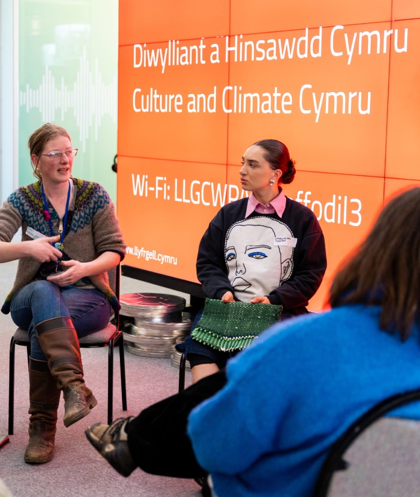 Two women sit and talk in front of an orange wall displaying Culture and Climate Cymru and Wi-Fi details. One woman gestures while the other listens, holding a green bag.