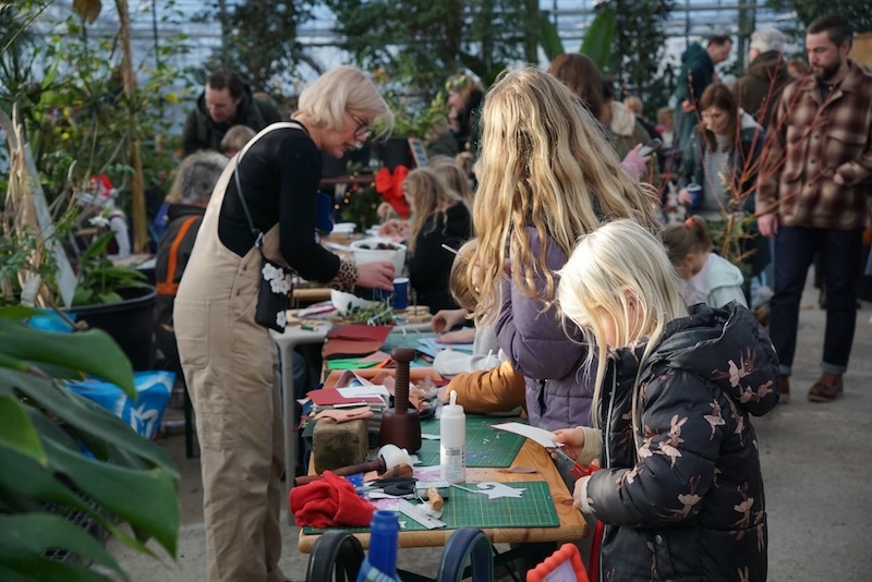 People of various ages participate in an arts and crafts activity at long tables in a busy indoor space filled with plants.