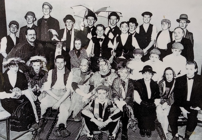 A group of men and women in vintage costumes, hats, and vests pose together for a black-and-white group photo, seated and standing in several rows.