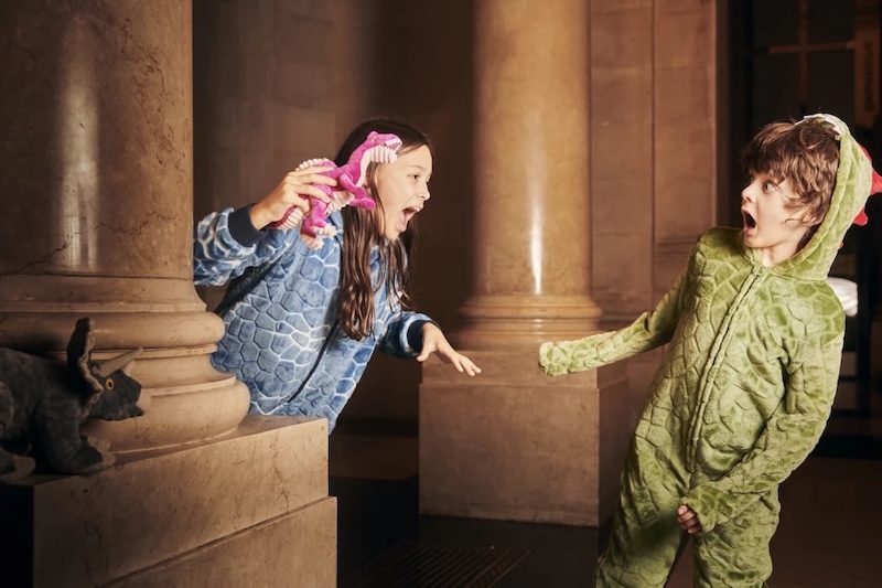 Two children in dinosaur costumes playfully scare each other near large stone columns indoors.