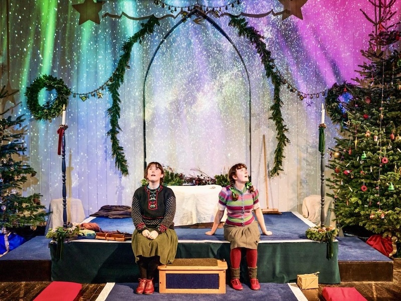 Two people sit on a stage decorated with wreaths, Christmas trees, and festive lights, looking upward under an arch and colorful backdrop.