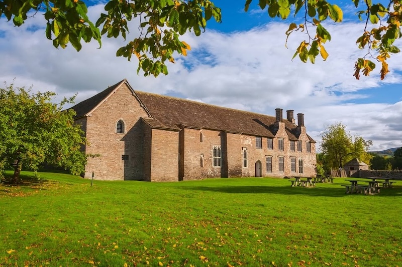 Large historic stone building with multiple chimneys, set on a grassy lawn with picnic tables and trees under a partly cloudy sky.
