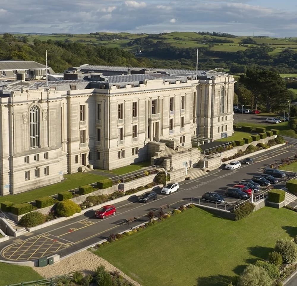A large stone building with columns and arched windows sits beside a parking lot, surrounded by greenery and hills under a partly cloudy sky.