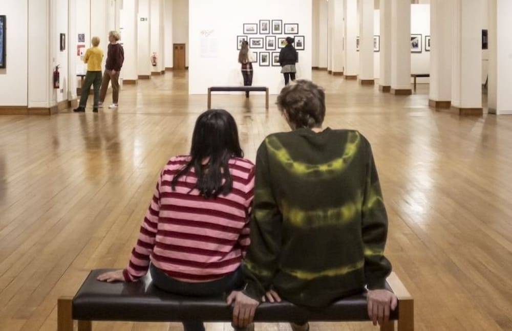Two people sit on a bench in an art gallery, facing a wall of framed photos, with several other visitors walking and viewing the exhibits.