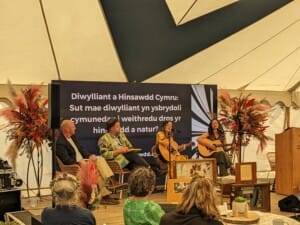Two women sitting on a stage playing guitar and singing, with other people watching.